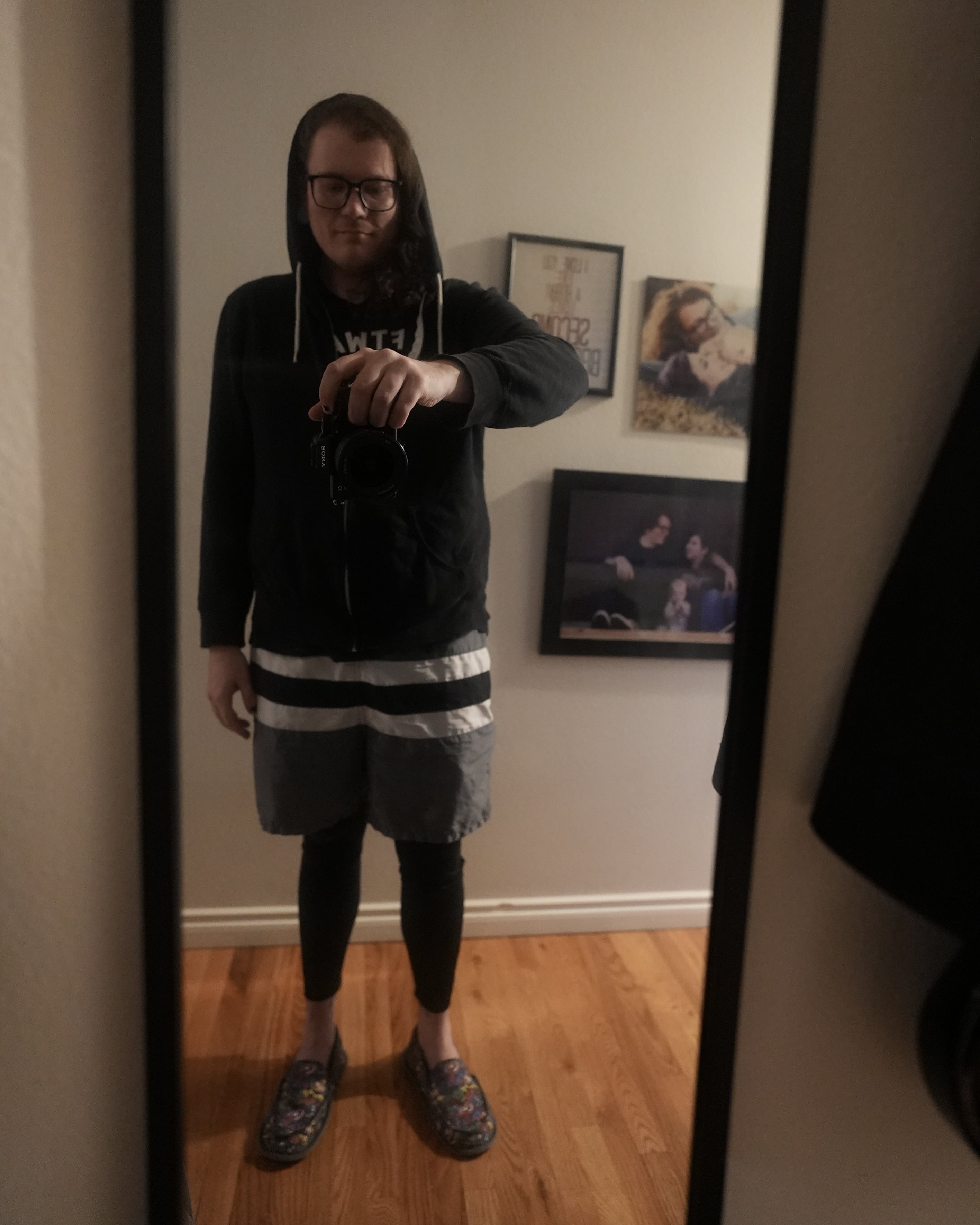 Me standing in a mirror taking a selfie. I'm' wearing black excercise tights under swimshorts and a hoodie and my comic book slippers. You can see blurry photos of our family on the wall behind me.